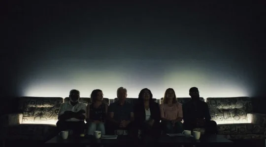 Un groupe de silhouettes assises côte à côte dans une salle sombre, réunies autour de la lueur chaleureuse d'une projection culturelle partagée